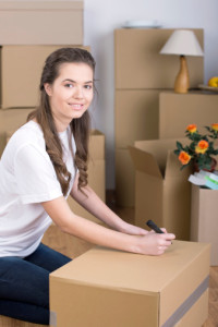 Apartments in The Woodlands For Rent A woman kneels and labels a cardboard box with a marker, preparing to move to her new apartment. Behind her, several other boxes, a lamp, and a vase with flowers await. She is wearing a white shirt and jeans. The Woodlands Lodge Apartments 2500 S Millbend Dr, The Woodlands, TX 77380