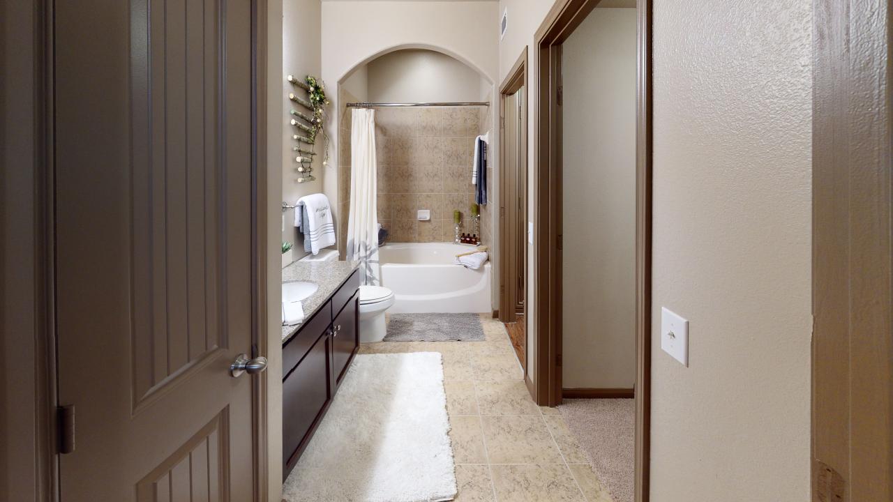 Apartments in The Woodlands For Rent A narrow bathroom in one bedroom apartments in The Woodlands features a white bathtub with a tile surround, a long counter with a sink on the left, and a floor covered with a light rug and tiles. A towel rack is mounted on the wall. The Woodlands Lodge Apartments 2500 S Millbend Dr, The Woodlands, TX 77380