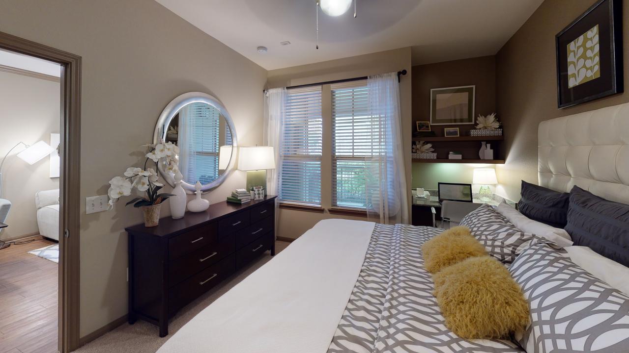 Apartments in The Woodlands For Rent A cozy one-bedroom apartment in The Woodlands features a bedroom with a bed, patterned bedding, fluffy pillows, a dresser with a round mirror, a window adorned with blinds and sheer curtains, and even includes a small workspace with a desk and computer. The Woodlands Lodge Apartments 2500 S Millbend Dr, The Woodlands, TX 77380