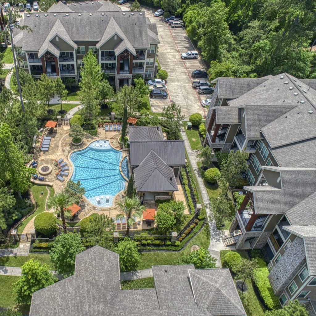 Apartments in The Woodlands For Rent Aerial view of an apartment complex featuring two multi-story buildings with one-bedroom apartments in The Woodlands, a swimming pool with lounge chairs, a hot tub, and a small central building, surrounded by greenery and parked cars. The Woodlands Lodge Apartments 2500 S Millbend Dr, The Woodlands, TX 77380