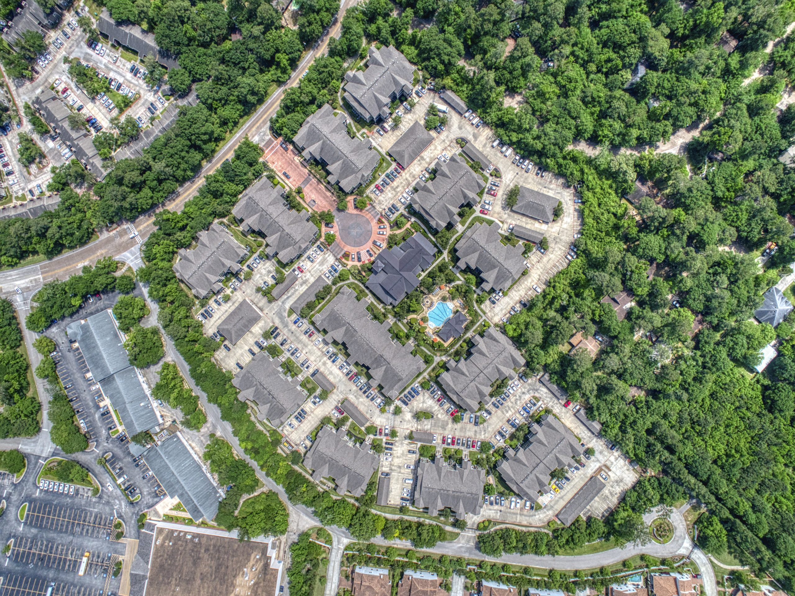 Apartments in The Woodlands For Rent Aerial view of a residential complex with multiple apartment buildings, a central circular plaza, a pool, and surrounded by dense greenery and parking lots. The Woodlands Lodge Apartments 2500 S Millbend Dr, The Woodlands, TX 77380