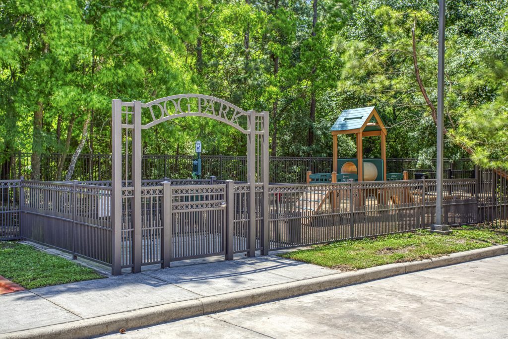 Apartments in The Woodlands For Rent Small dog park with a fenced area, including a small playground structure with a roof. Green trees and bushes surround the park. A paved pathway leads to the entrance gate labeled "Dog Park". The Woodlands Lodge Apartments 2500 S Millbend Dr, The Woodlands, TX 77380