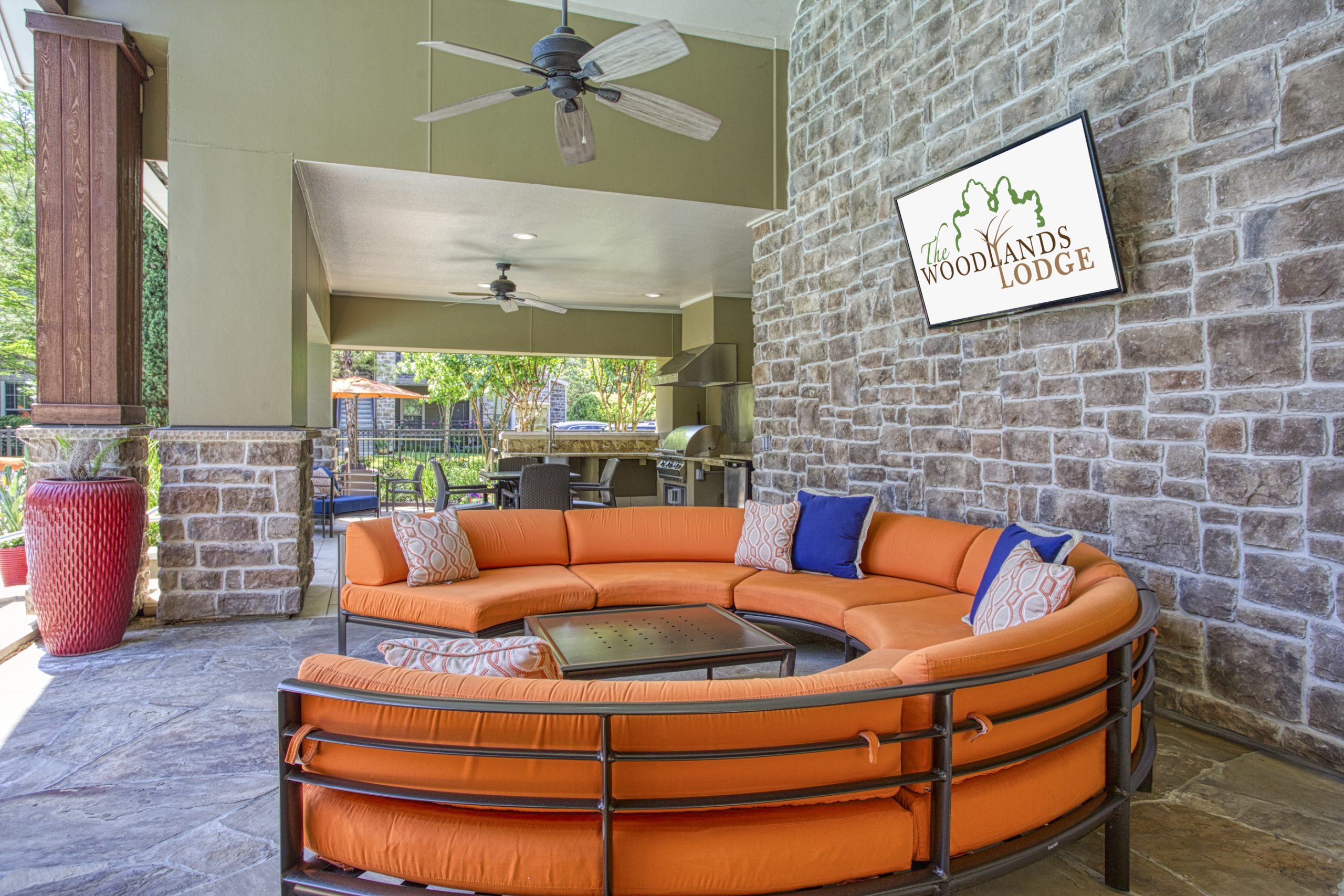 Apartments in The Woodlands For Rent Outdoor lounge area with a circular orange sofa, a ceiling fan, and a mounted TV displaying "The Woodlands Lodge" logo. The background features a stone wall and an outdoor grill, perfectly capturing the upscale ambiance of Apartments In The Woodlands. The Woodlands Lodge Apartments 2500 S Millbend Dr, The Woodlands, TX 77380