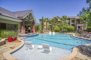 Apartments in The Woodlands For Rent Image of a resort-style outdoor pool area with lounge chairs, a covered seating area, and landscaped surroundings. Multiple buildings are visible in the background under a clear blue sky. The Woodlands Lodge Apartments 2500 S Millbend Dr, The Woodlands, TX 77380