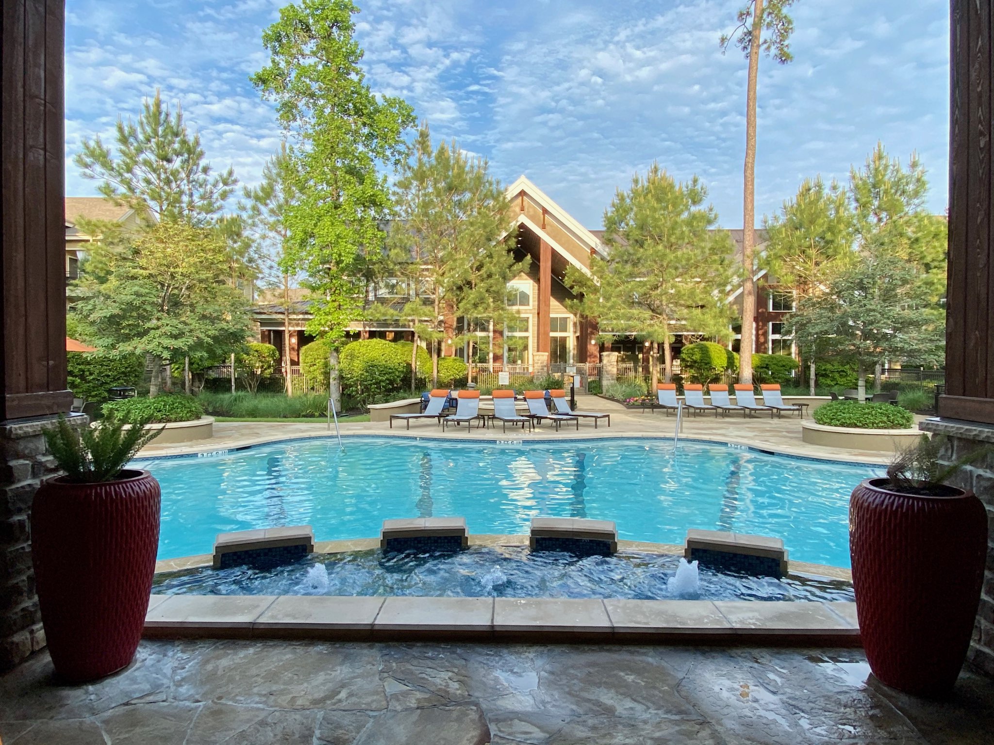 Apartments in The Woodlands For Rent Outdoor pool area with surrounding lounge chairs, landscaped greenery, and a clear blue sky. Scene viewed from a shaded patio with potted plants. The Woodlands Lodge Apartments 2500 S Millbend Dr, The Woodlands, TX 77380